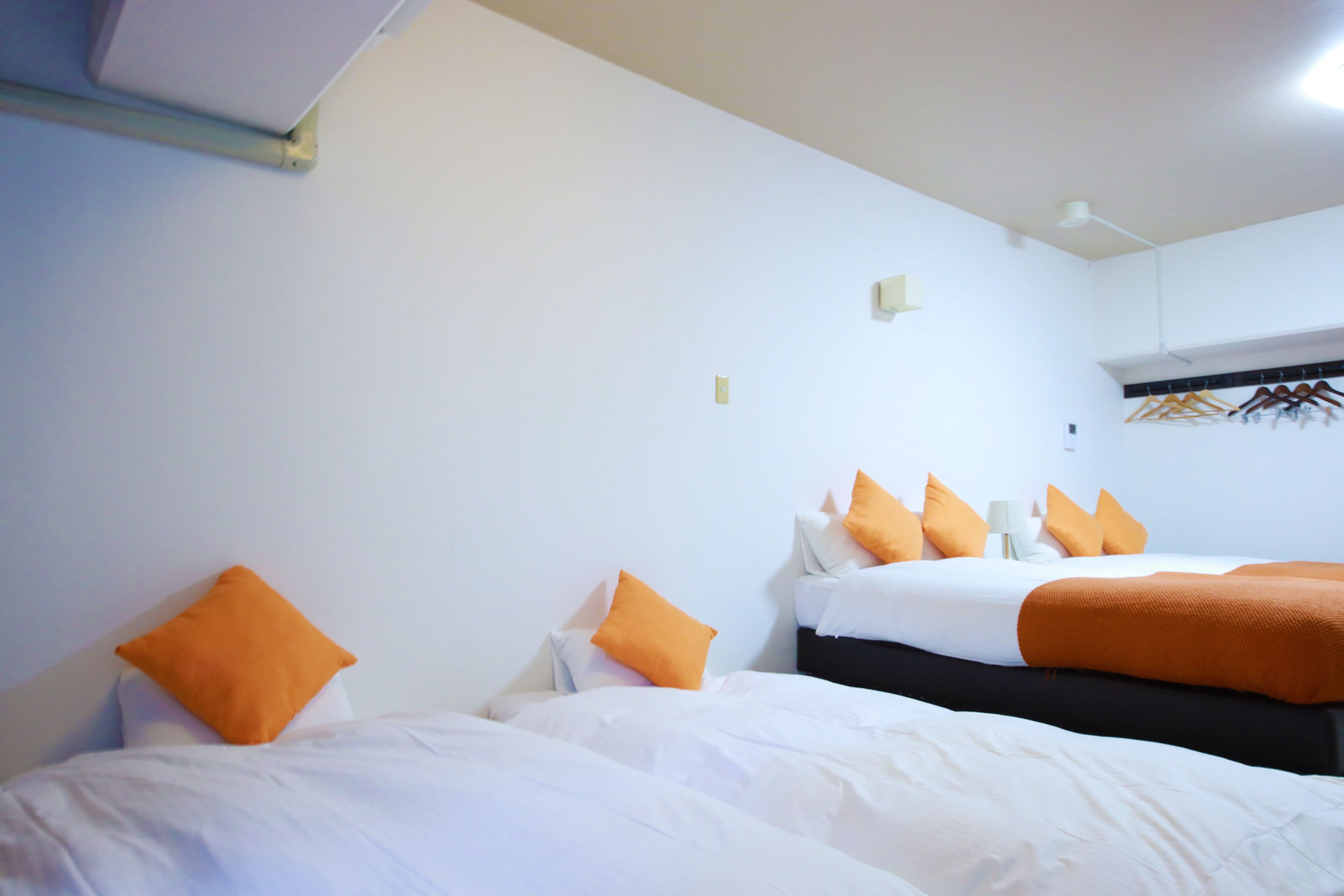 A hotel room with two beds, each with white bedding and orange pillows. There is a wall-mounted coat rack with hangers.