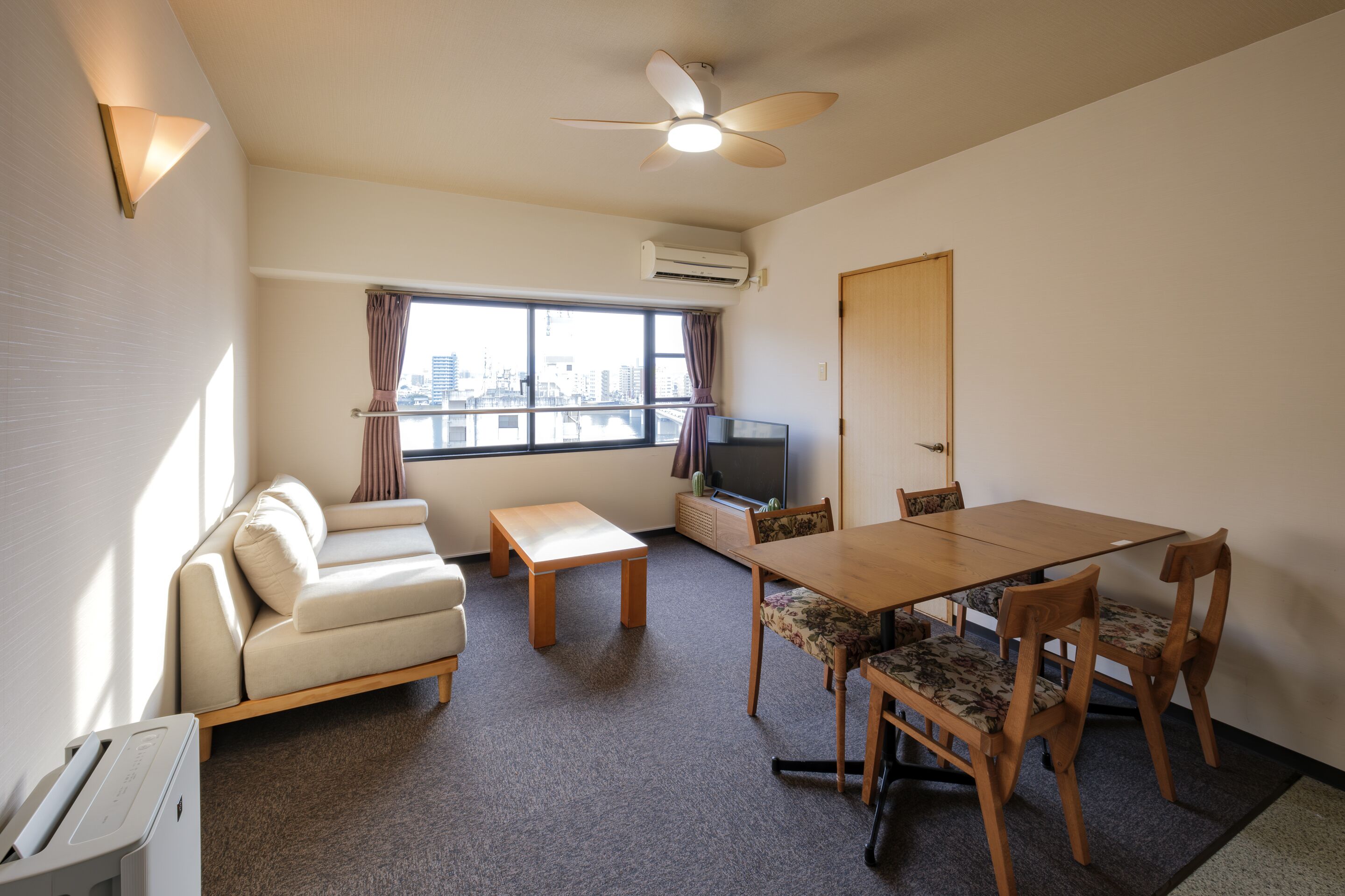 Un espacio habitable compacto con sofá, una mesa de comedor con sillas, ventilador de techo, televisor y ventana con vista a la ciudad.