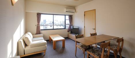 A compact living space with a sofa, a dining table with chairs, a ceiling fan, a television, and a window with a city view.