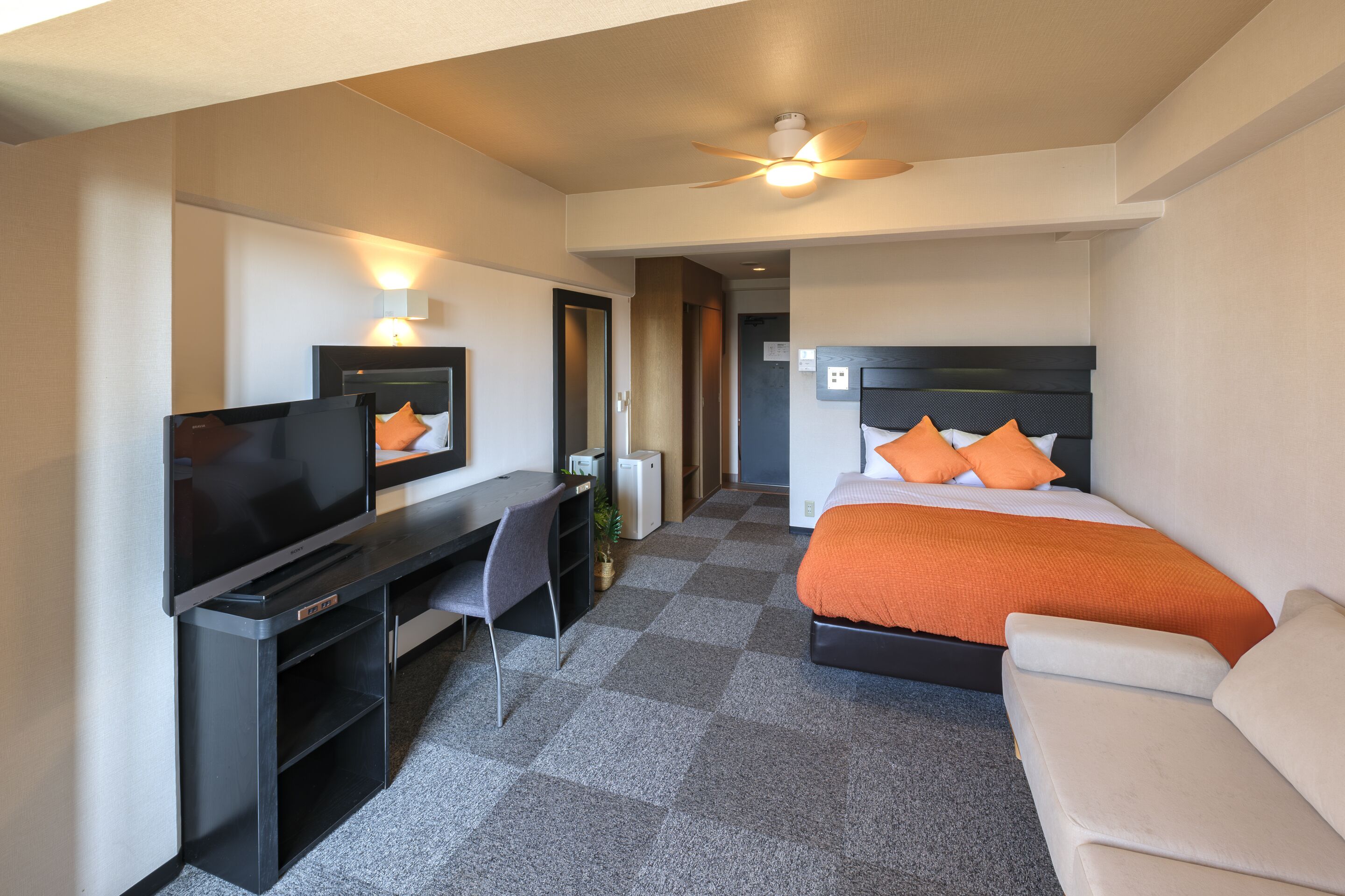 Ein Hotelzimmer mit Bett, Schreibtisch, Flachbildfernseher, Deckenventilator und Sofa.