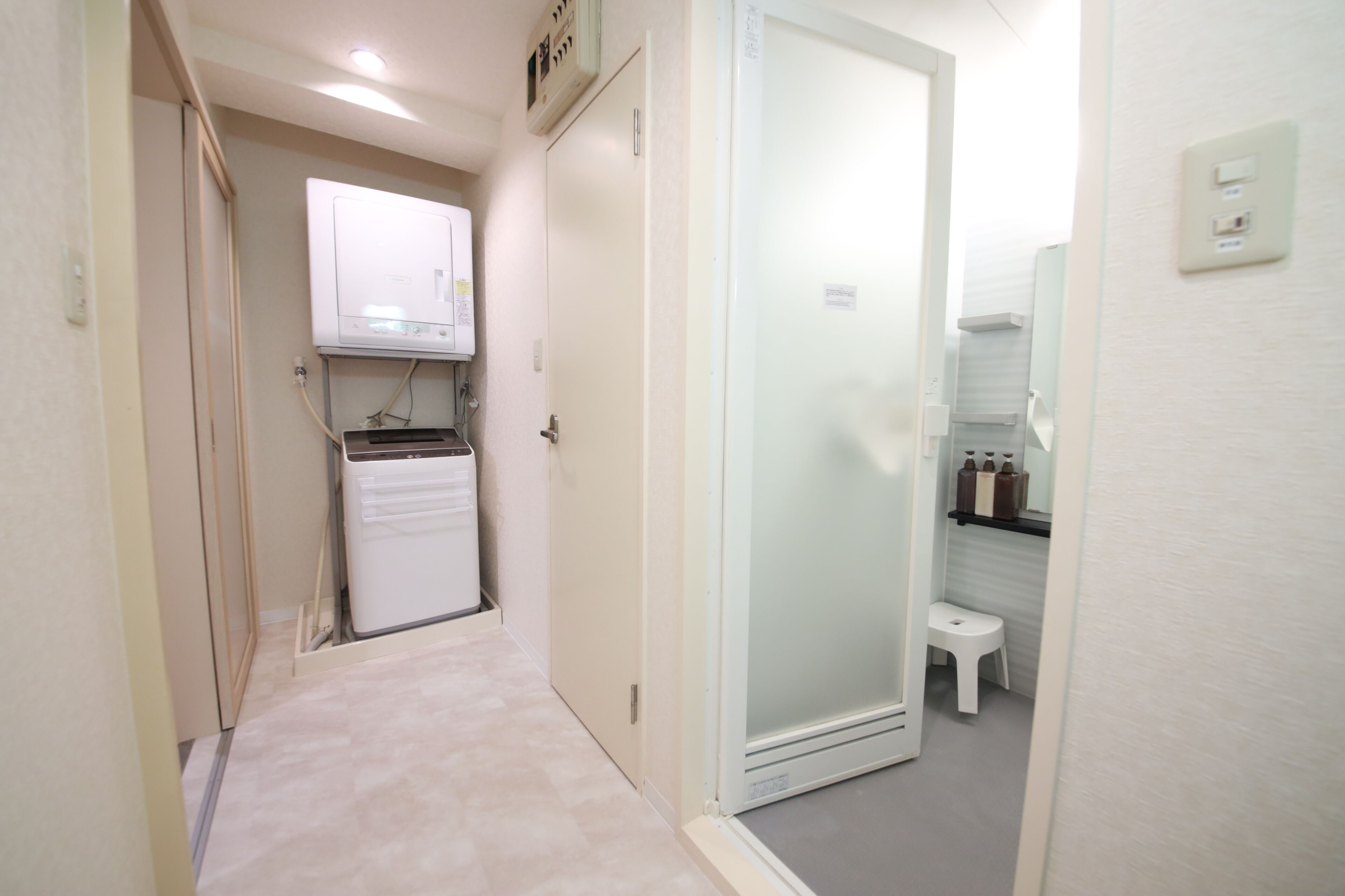 Un pasillo estrecho con un refrigerador blanco, una puerta de vidrio que da a un baño y un taburete pequeño.