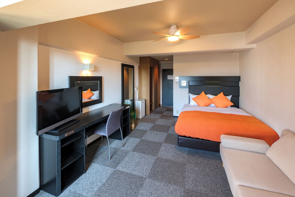Ett hotellrum med en säng, ett skrivbord med en platt-TV, en takfläkt och en soffa.