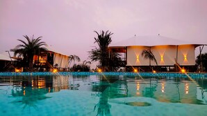 A resort pool area with a clear view of the pool, palm trees, and a building with a curved roof and illuminated walls. - ShriGo Resort Pushkar (Peesangan)