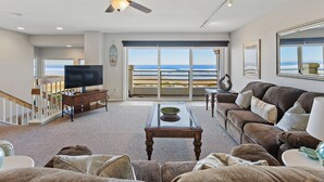 Un salon spacieux doté d’un grand téléviseur, de sièges confortables, d’un ventilateur de plafond et offrant une vue sur l’océan grâce à de grandes fenêtres.