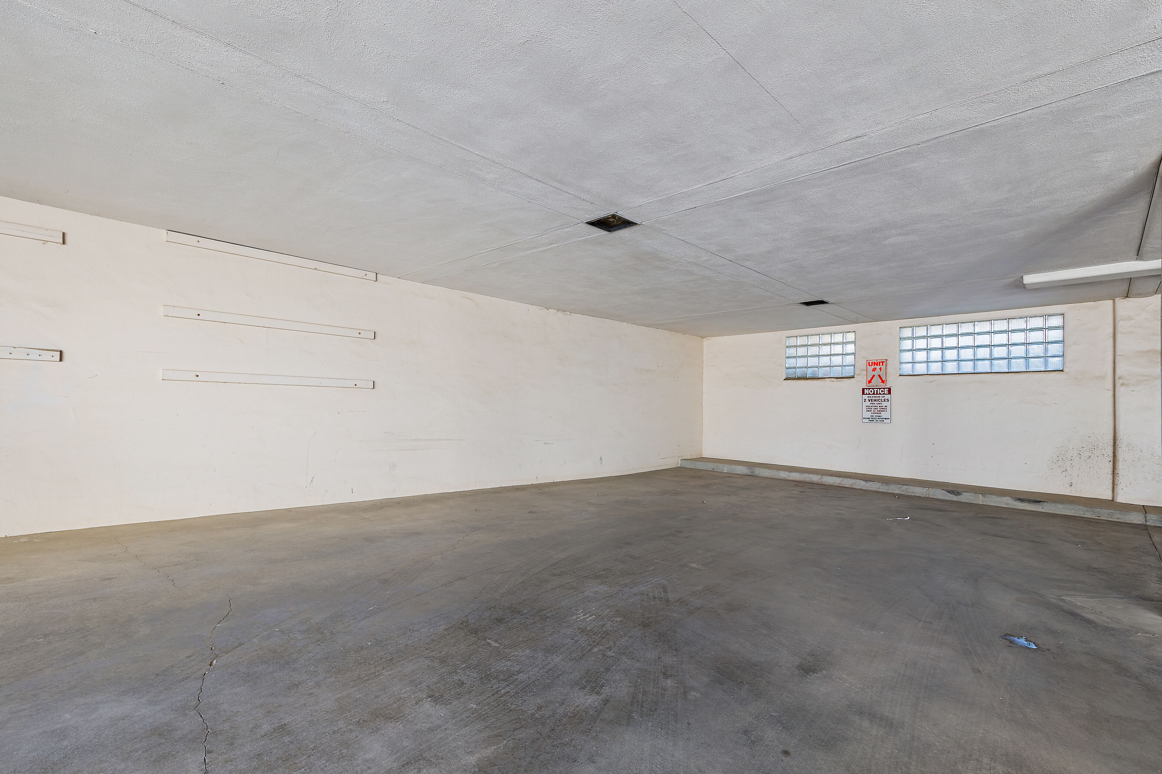 Una habitación amplia y vacía con piso de concreto y paredes blancas. Hay dos ventanas con rejas, un letrero en la pared e iluminación empotrada en el techo.