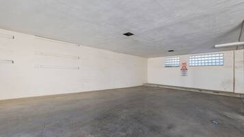 Una habitación amplia y vacía con piso de concreto y paredes blancas. Hay dos ventanas con rejas, un letrero en la pared e iluminación empotrada en el techo.