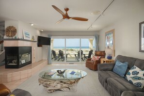 A living room with a glass coffee table, a fireplace, a ceiling fan, and a view of the beach through large sliding doors.