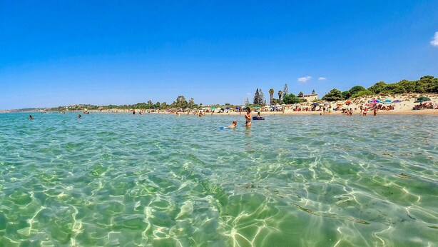 Une plage avec une eau turquoise et limpide, des gens qui se baignent et prennent des bains de soleil, et des parasols de plage.