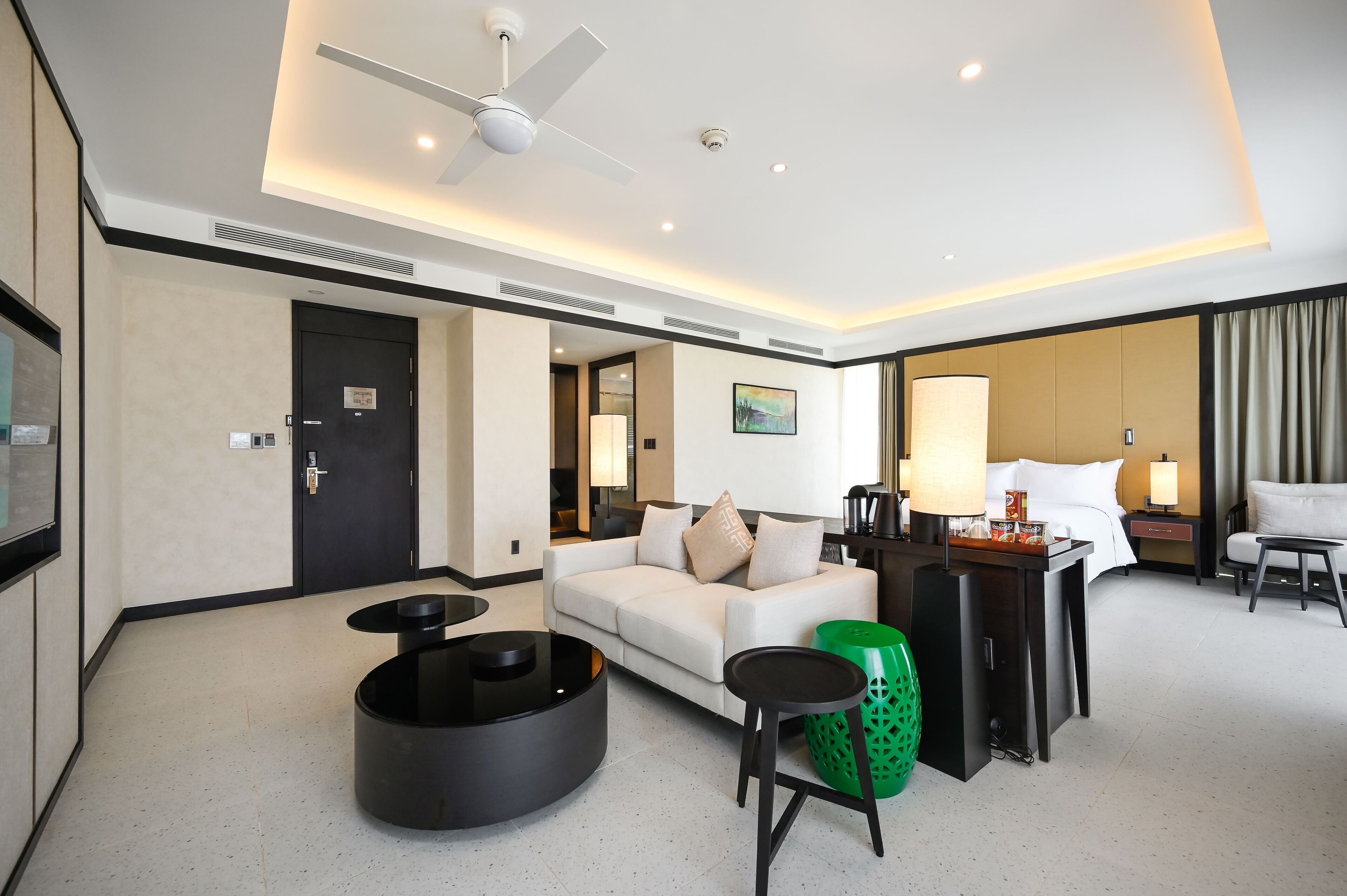 A modern hotel room with a white sofa, a round black coffee table, a green trash bin, a desk with a lamp, and a bed with a white headboard.