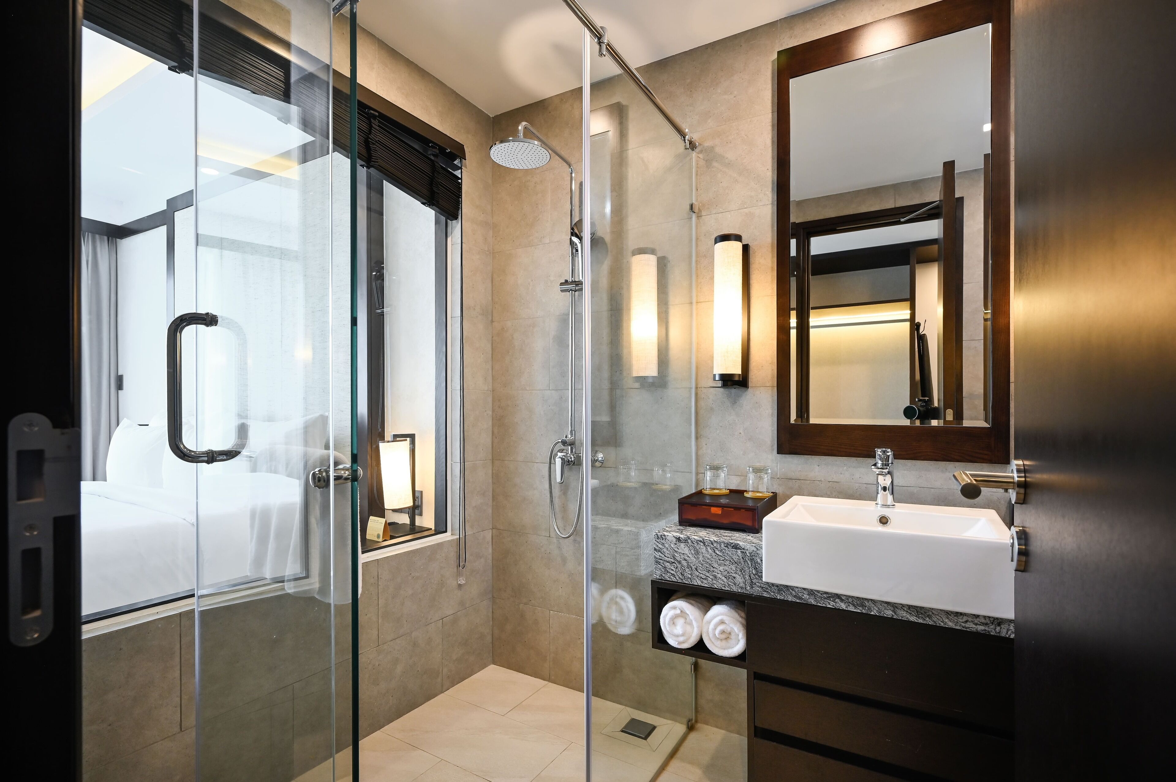Un baño de hotel moderno con ducha envuelta en vidrio, un espejo grande, un lavamanos con encimera de mármol y una mesita de noche con lámpara.