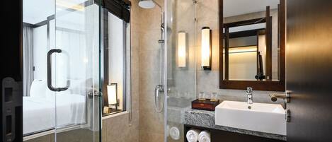 Un baño de hotel moderno con ducha envuelta en vidrio, un espejo grande, un lavamanos con encimera de mármol y una mesita de noche con lámpara.