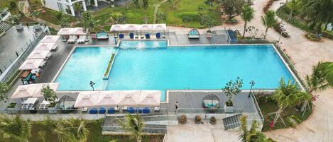 A large rectangular swimming pool with multiple lounge areas and umbrellas, surrounded by palm trees and a landscaped garden.