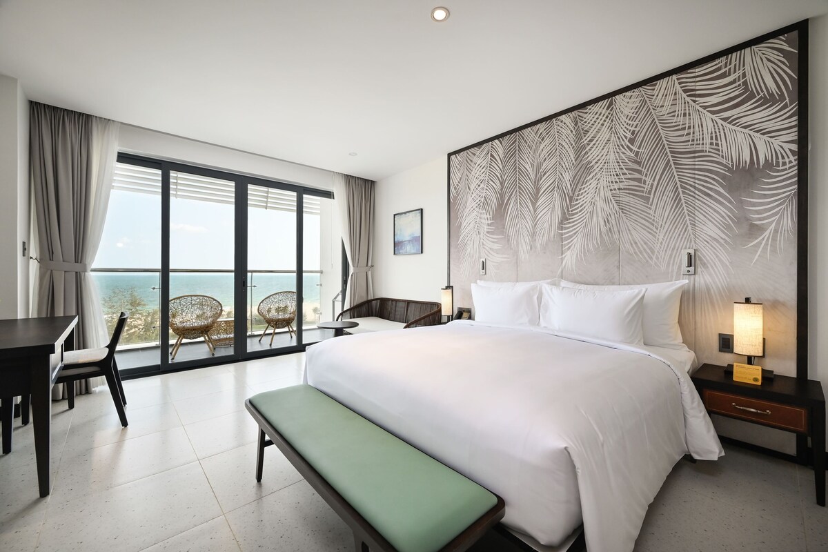 A hotel room with a large bed, a bench, a desk, and a chair. There is a large window with a view of the ocean.
