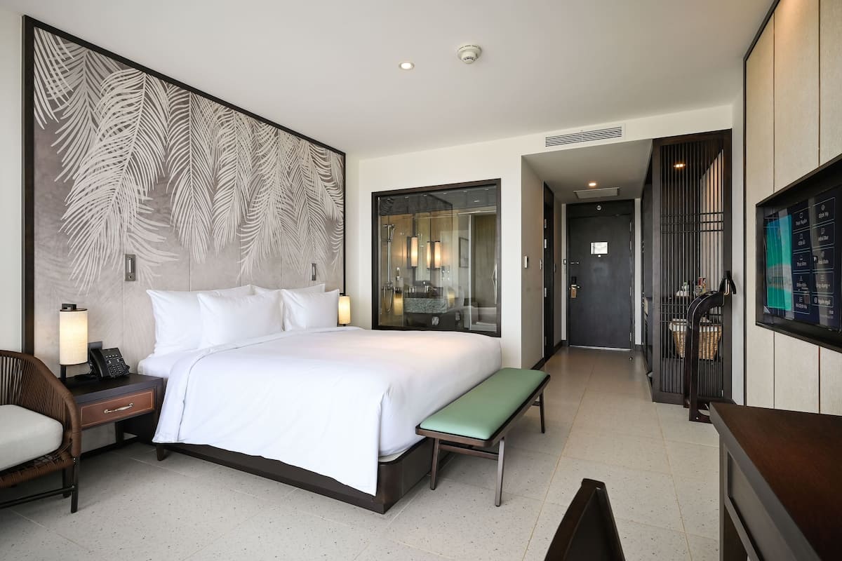 A hotel room with a large bed, bedside table with a lamp, a bench, a TV, and a bathroom visible through a glass door.