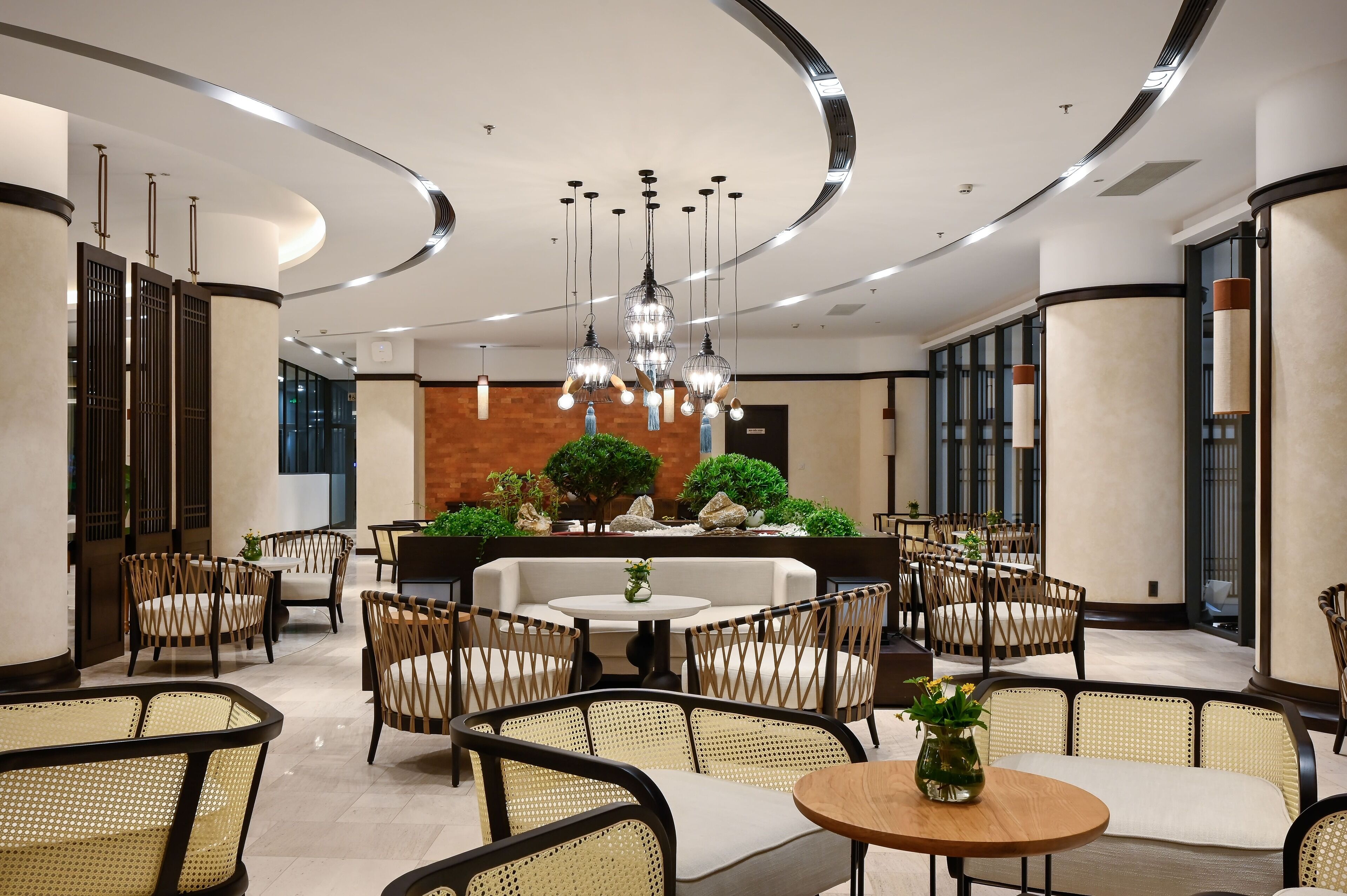 Un hall d'hôtel moderne avec des tables rondes, des fauteuils en osier et un espace salon central agrémenté d'un canapé blanc.