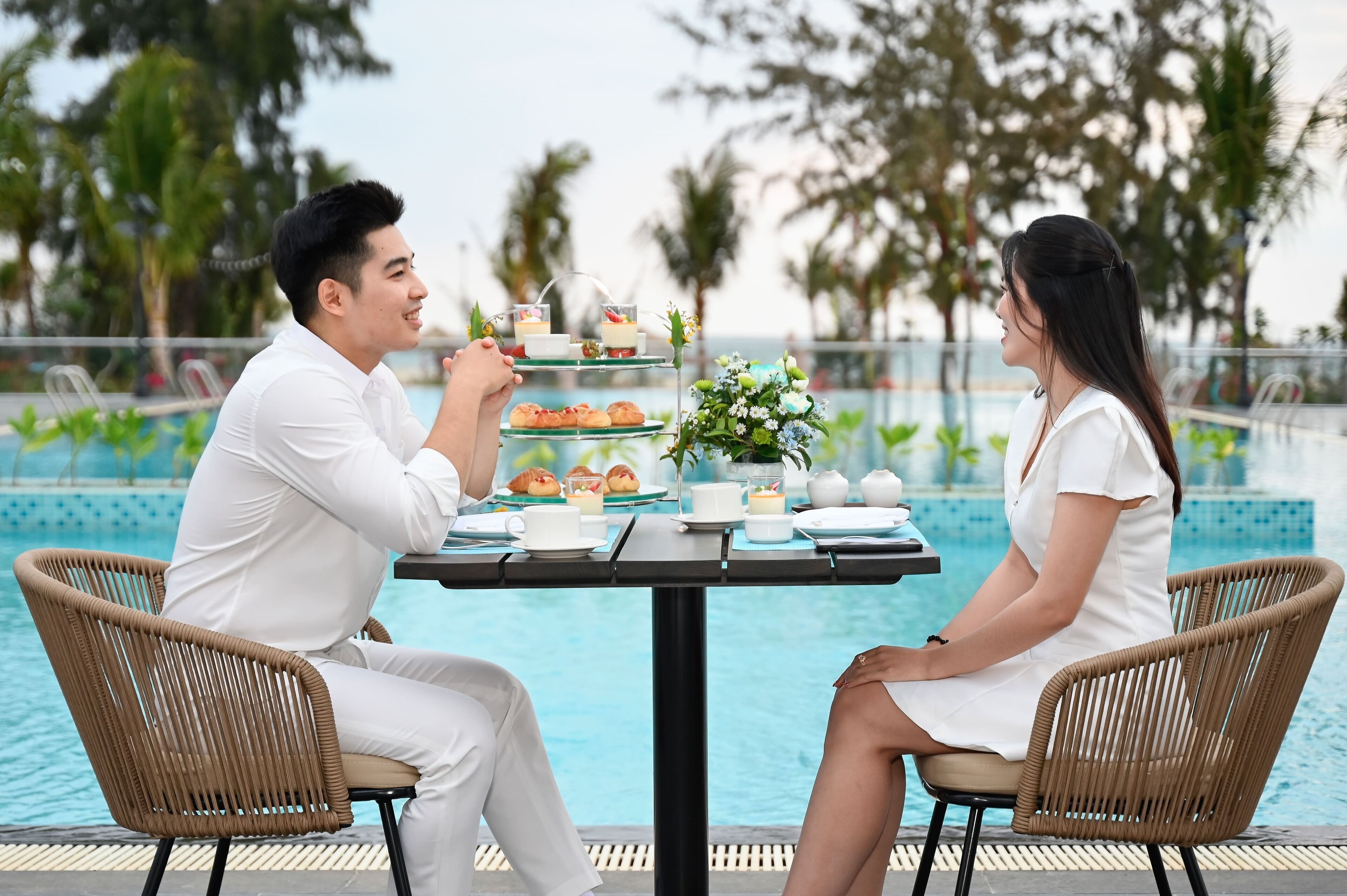 Una mesa de comedor con un expositor de postres de tres niveles, dos personas sentadas y una piscina al fondo.