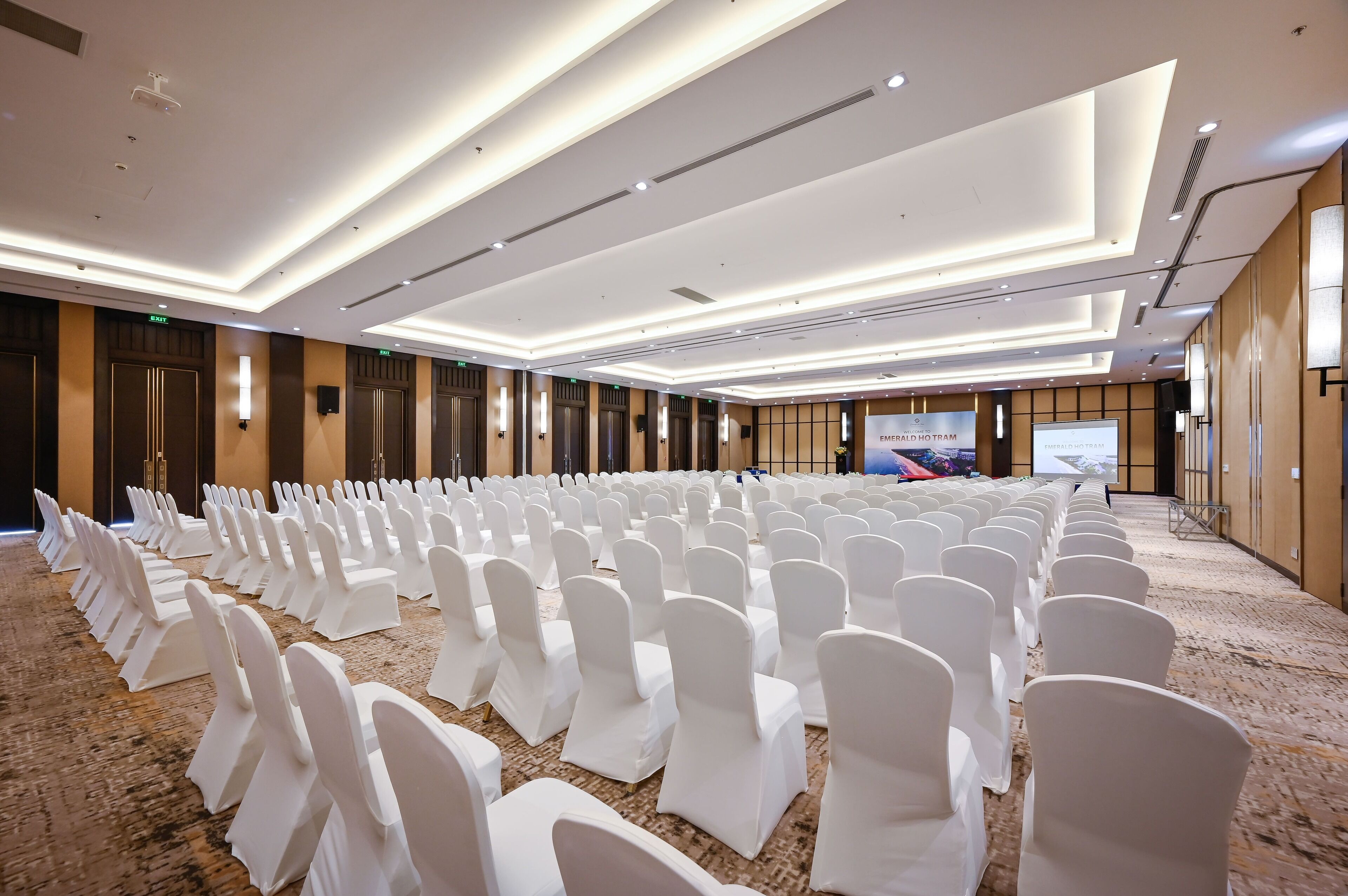 Un salón de conferencias con hileras de sillas blancas, piso alfombrado y un diseño de techo moderno.