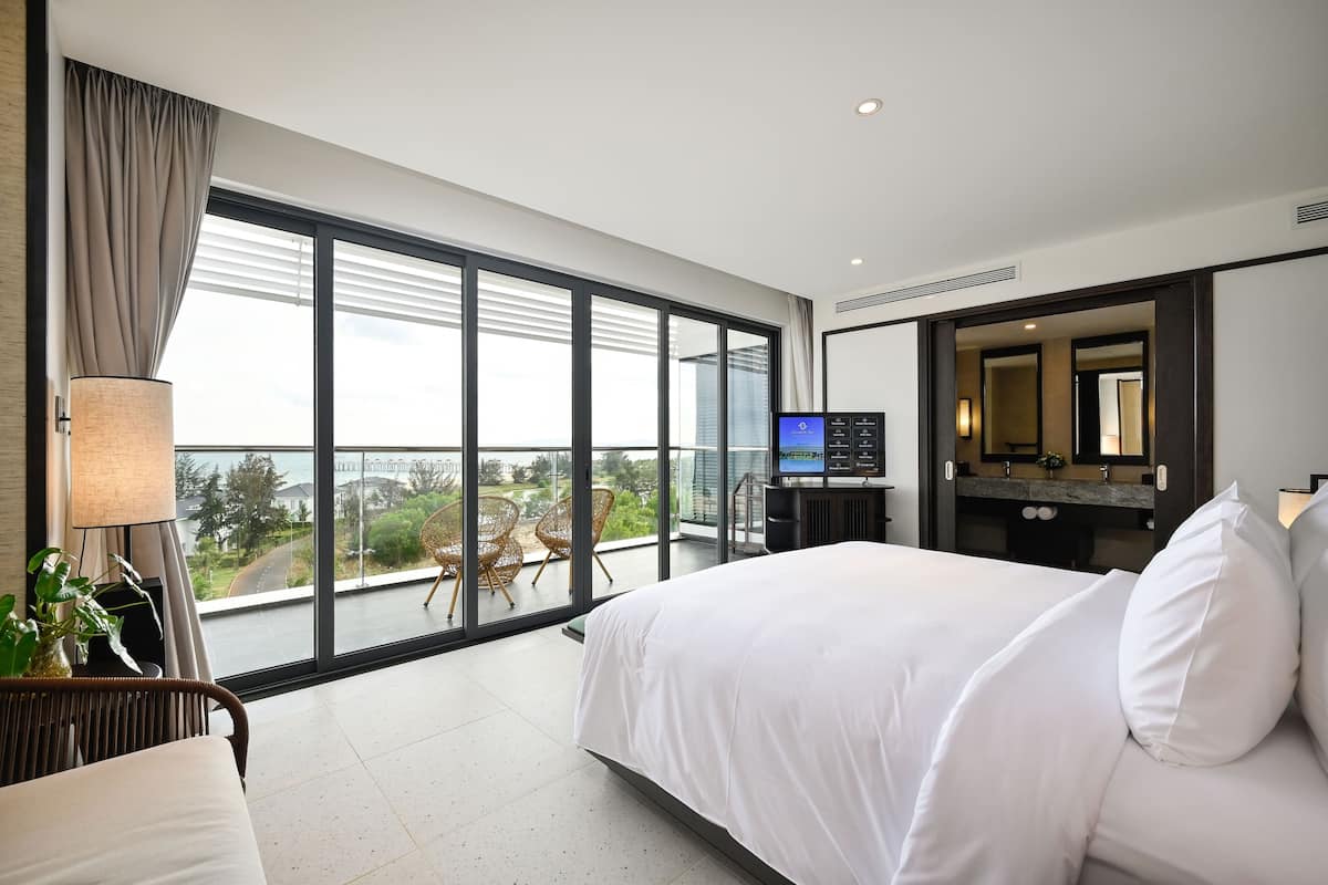 A modern hotel room with a large bed, a television, a bathroom with a mirror, and a balcony with a view of the city.