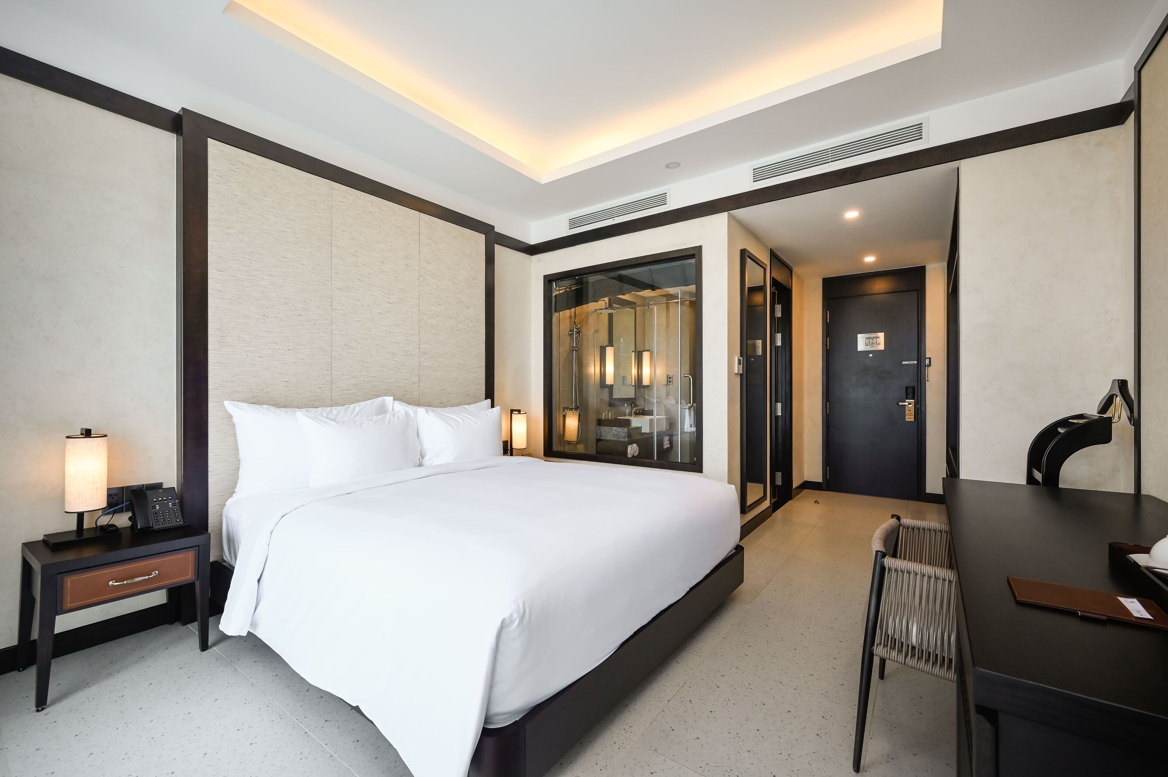 Habitación de hotel con una cama grande, mesita de noche con lámpara, escritorio con silla y zona de ducha con mampara de vidrio.