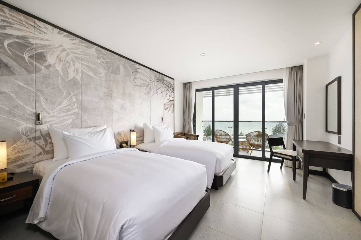 A hotel room with two beds, a desk, and a view of the outdoors.