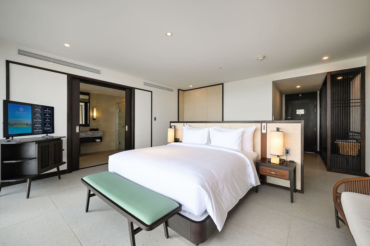 A hotel room with a bed, bedside tables, a desk, a bench, a television, and a bathroom visible through an open door.