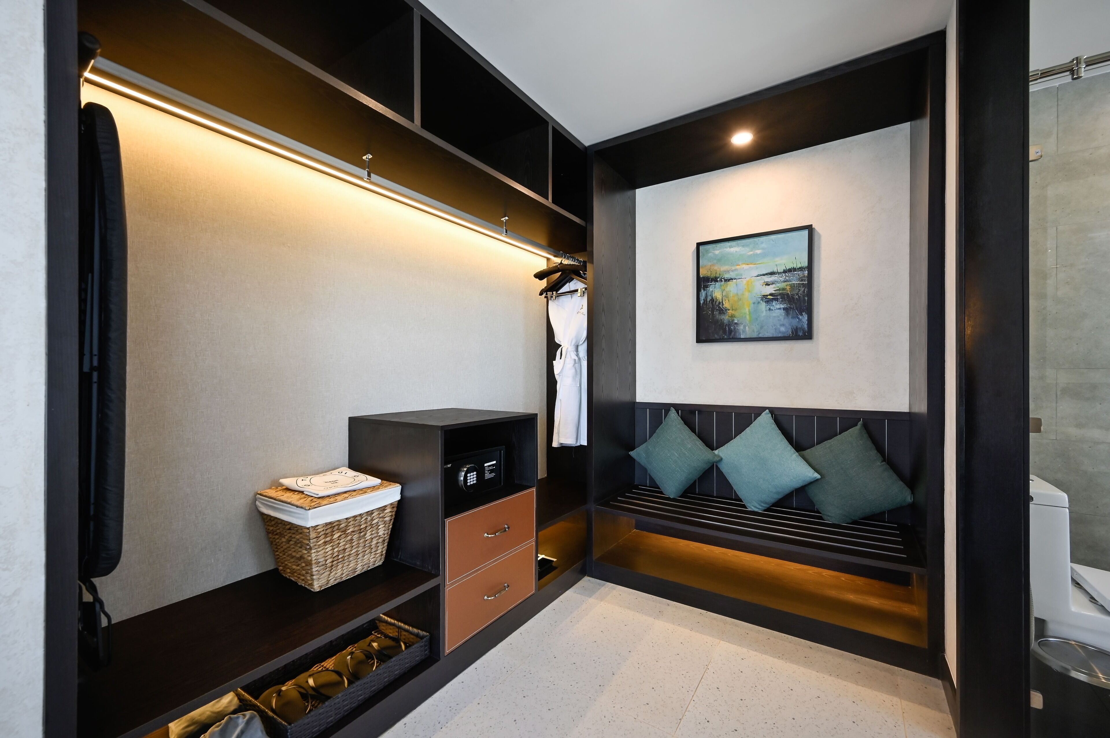 A modern hotel room with a built-in wardrobe, a seating area with cushions, a painting on the wall, and a small table with a basket.
