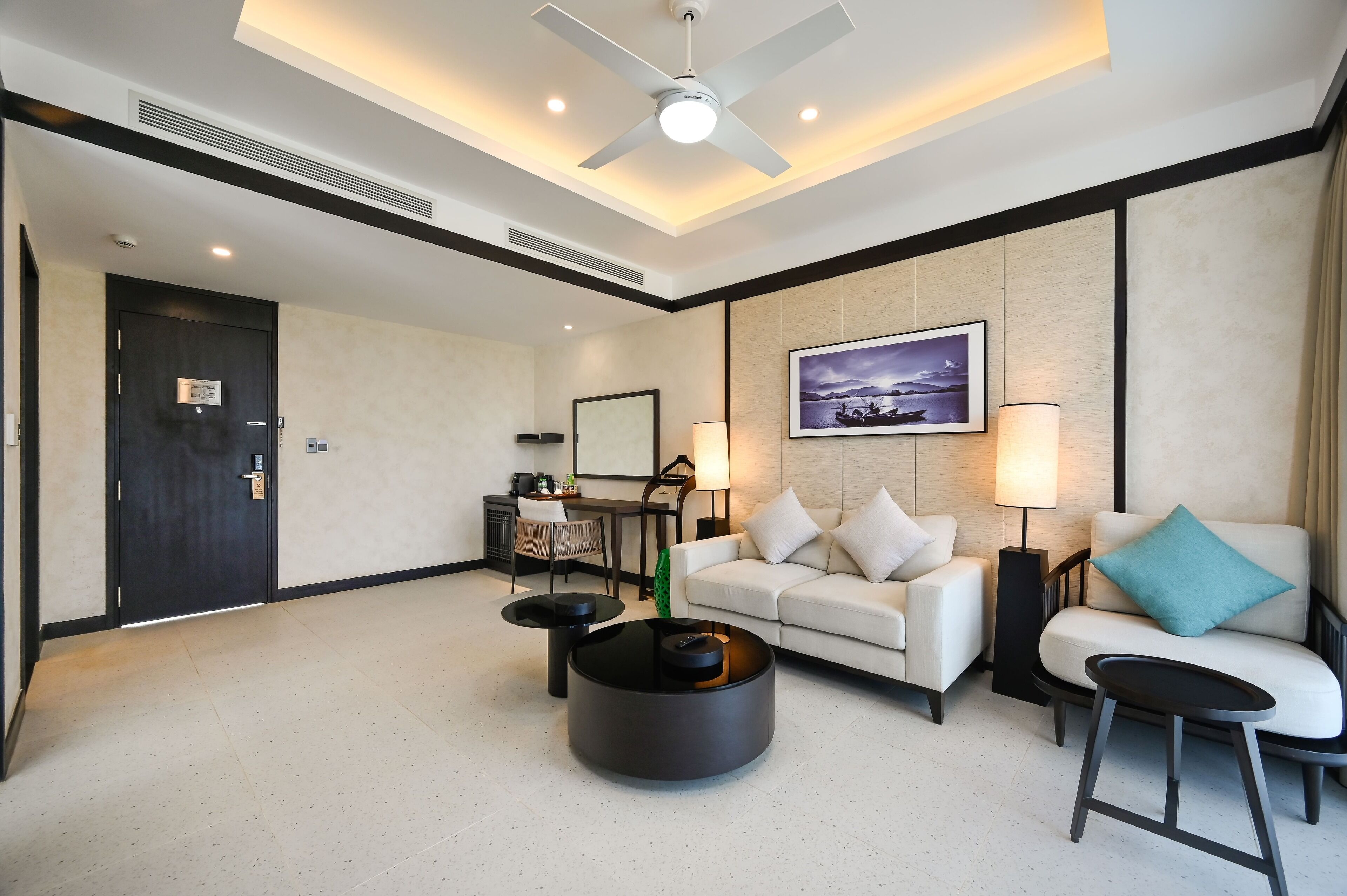 Une chambre d’hôtel moderne équipée d’un canapé, de deux tables d’appoint, d’une table basse, d’un ventilateur de plafond, d’un bureau avec une chaise, d’une lampe et d’un tableau encadré au mur.