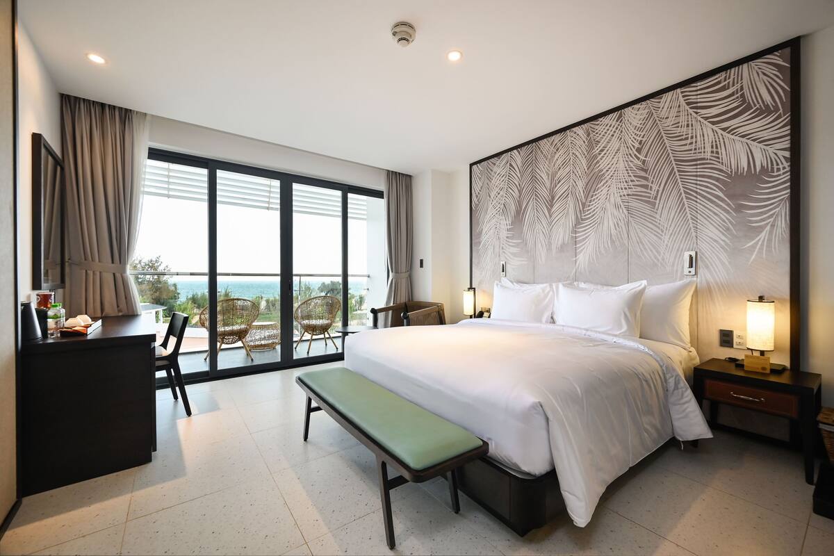 A modern hotel room with a large bed, a bench, a desk, and a view of the sea.