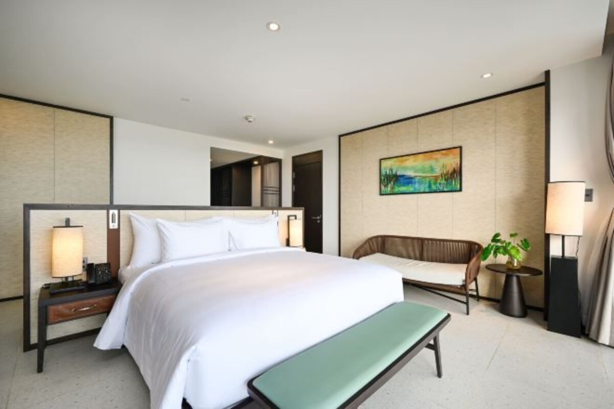 A hotel room with a large bed, a bench, a sofa, a desk with a lamp, and a painting on the wall.