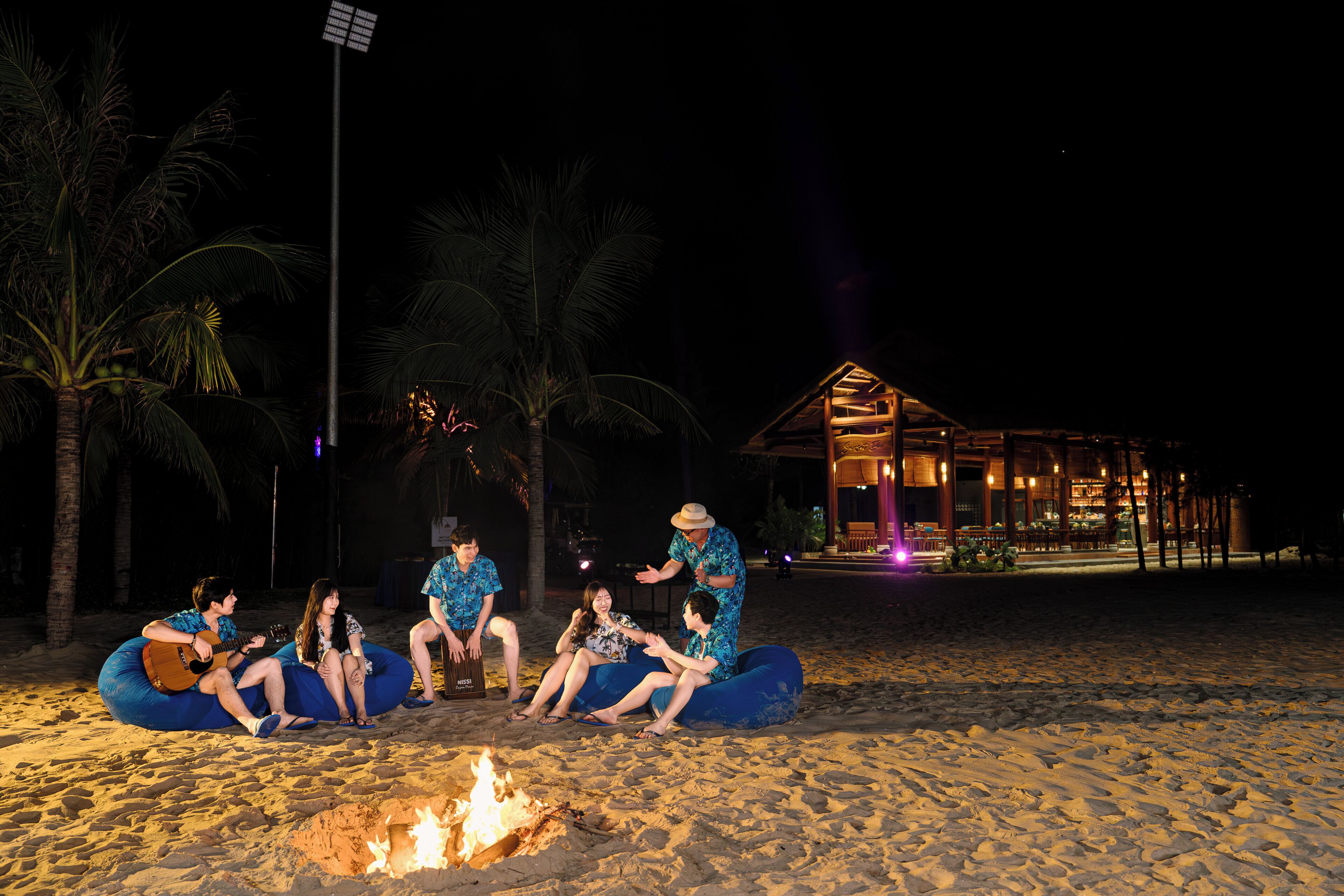 Eine Gruppe von Menschen sitzt bei Nacht am Lagerfeuer auf blauen Sitzsäcken an einem sandigen Strand, im Hintergrund ist ein Gebäude zu sehen.