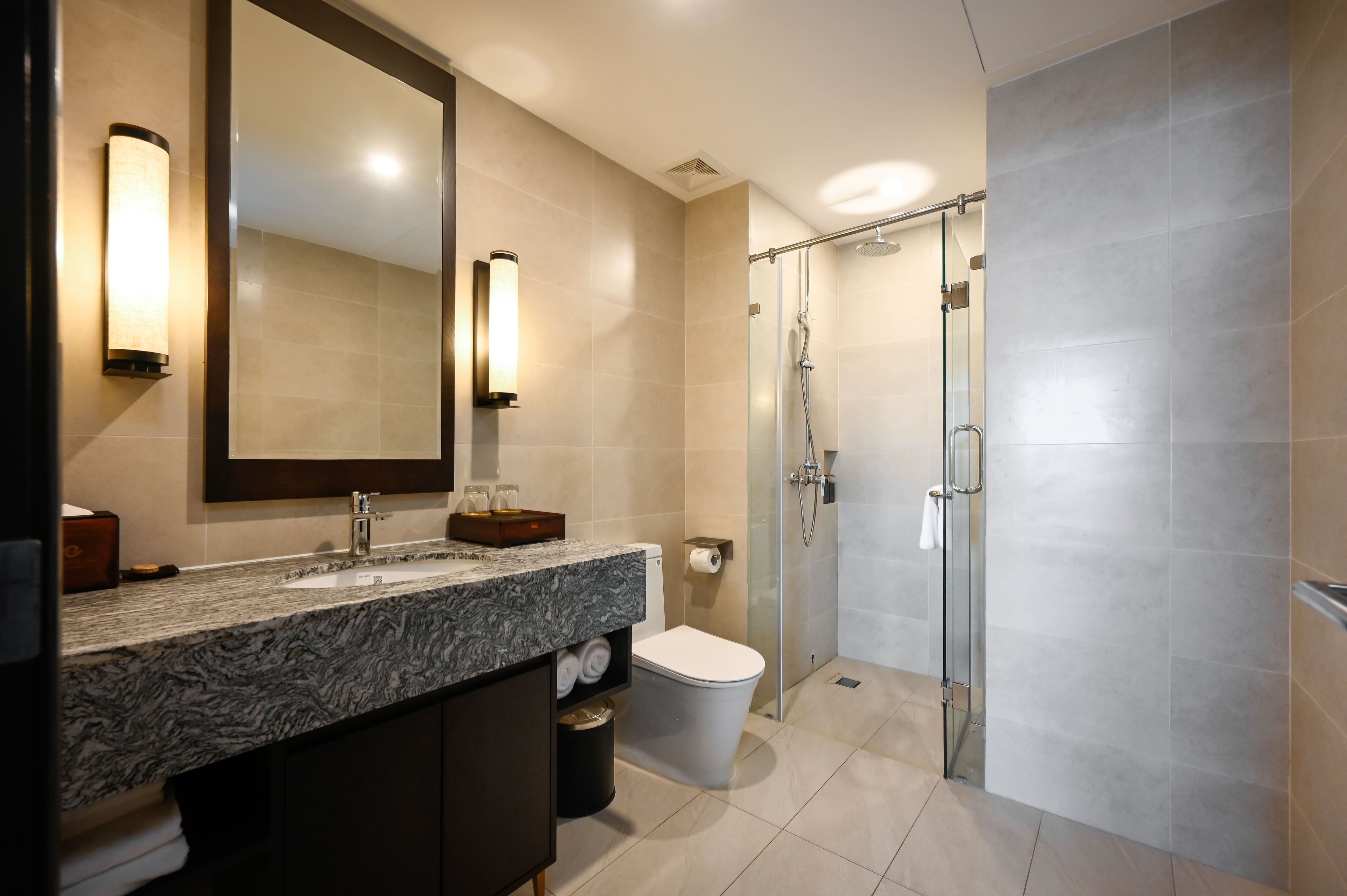 A modern bathroom with a granite countertop, a toilet, a glass shower enclosure, and a large mirror with wall sconces.