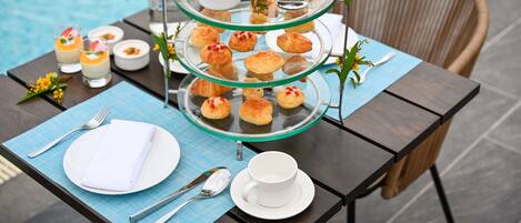 A three-tiered dessert tray with various pastries and desserts, a white cup, and a fork and spoon on a table by a pool.
