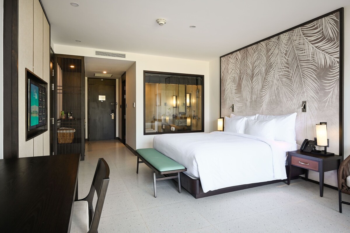 A hotel room with a large bed, a desk with a chair, a bench, a nightstand with a lamp, and a large mirror.