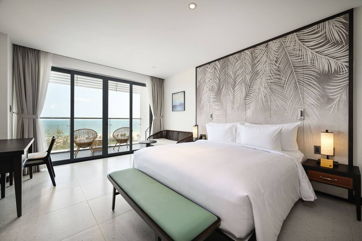 A hotel room with a large bed, a bench, a desk, and a chair. There is a large window with a view of the ocean.