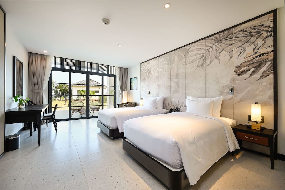A modern hotel room with two beds, a desk, and a large window offering a view of the outdoors.