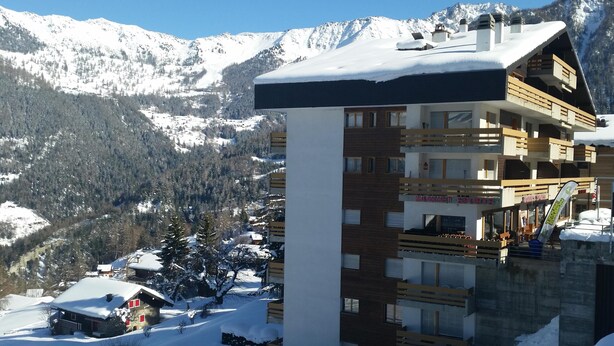 Een hotel met meerdere verdiepingen en balkons, een wintersportgebied op de achtergrond en besneeuwde bergen.