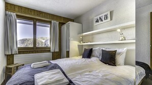 Een slaapkamer met een bed, twee kussens, een nachtkastje, een raam met gordijnen, een plank met een lamp en een lijst met een afbeelding aan de muur.