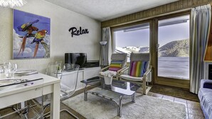 Een gezellige kamer met een eethoek, een televisie en een groot raam met uitzicht op een besneeuwde berg.