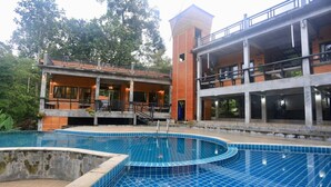 Un área de piscina con un edificio moderno al fondo, que presenta una estructura de dos pisos con balcones y una sección adicional que recuerda a una torre.