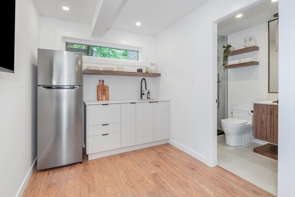 A modern kitchen with a stainless steel refrigerator, white cabinetry, wooden flooring, and a window above the sink.