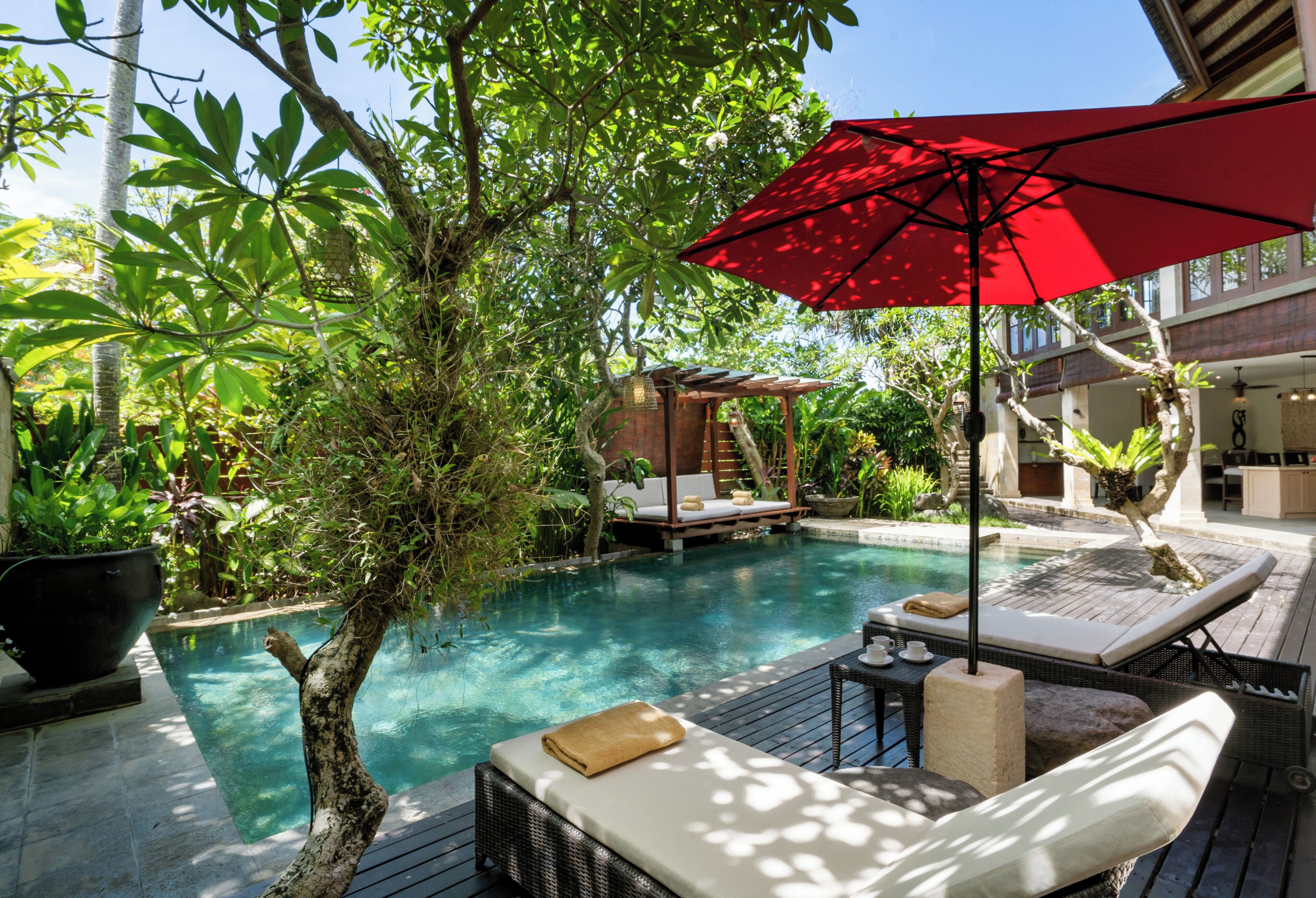 A poolside area with a lounge chair, a red umbrella, and a small seating area with a wooden structure.