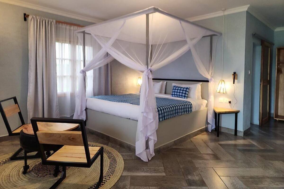 A bedroom with a four-poster canopy bed, a bedside table with a lamp, and a chair with a woven rug.
