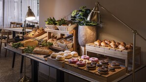 Una vitrina de panadería con panes y pasteles variados, mesas de madera y una planta grande.