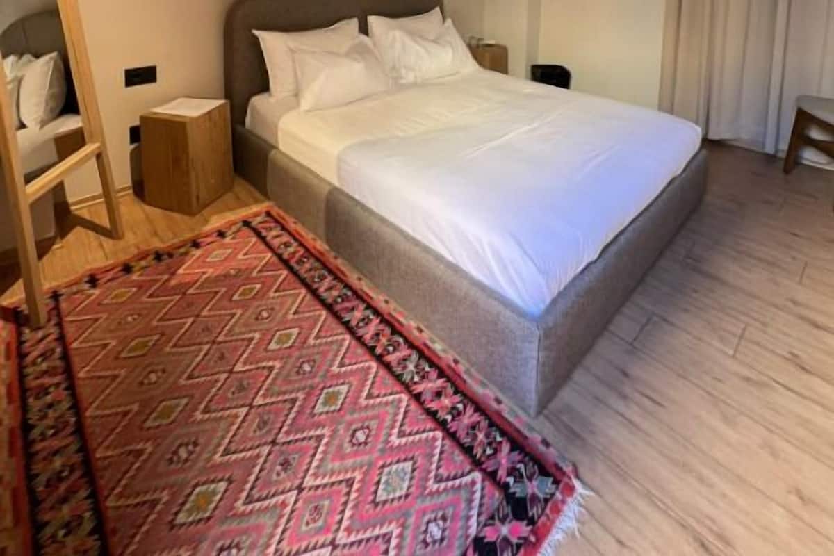Une chambre avec un plafond en bois, un lit avec une literie blanche, une petite table de chevet, une lampe de chevet, un tapis à motifs et une fenêtre avec des rideaux.