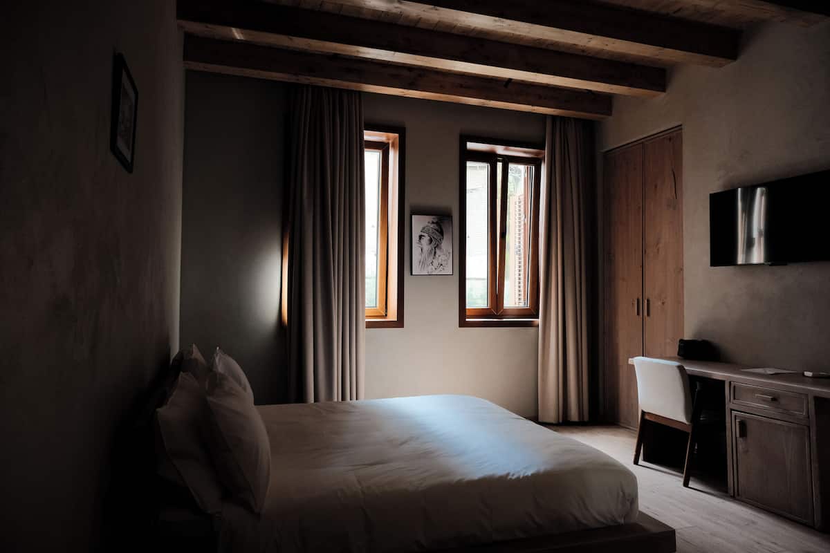 Une chambre avec un lit, un bureau, une chaise et un téléviseur. Il y a deux fenêtres avec des rideaux et un tableau accroché au mur.