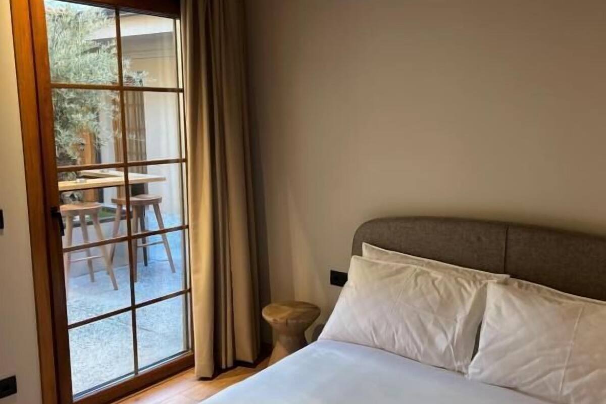 Une chambre dotée d’un grand lit, d’une tête de lit en bois, d’une petite table de chevet et d’une porte-fenêtre coulissante donnant sur un espace extérieur avec une table et des chaises.