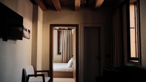 Un couloir étroit mène à une chambre qui contient un lit, une chaise, un téléviseur fixé au mur et une fenêtre avec des rideaux.