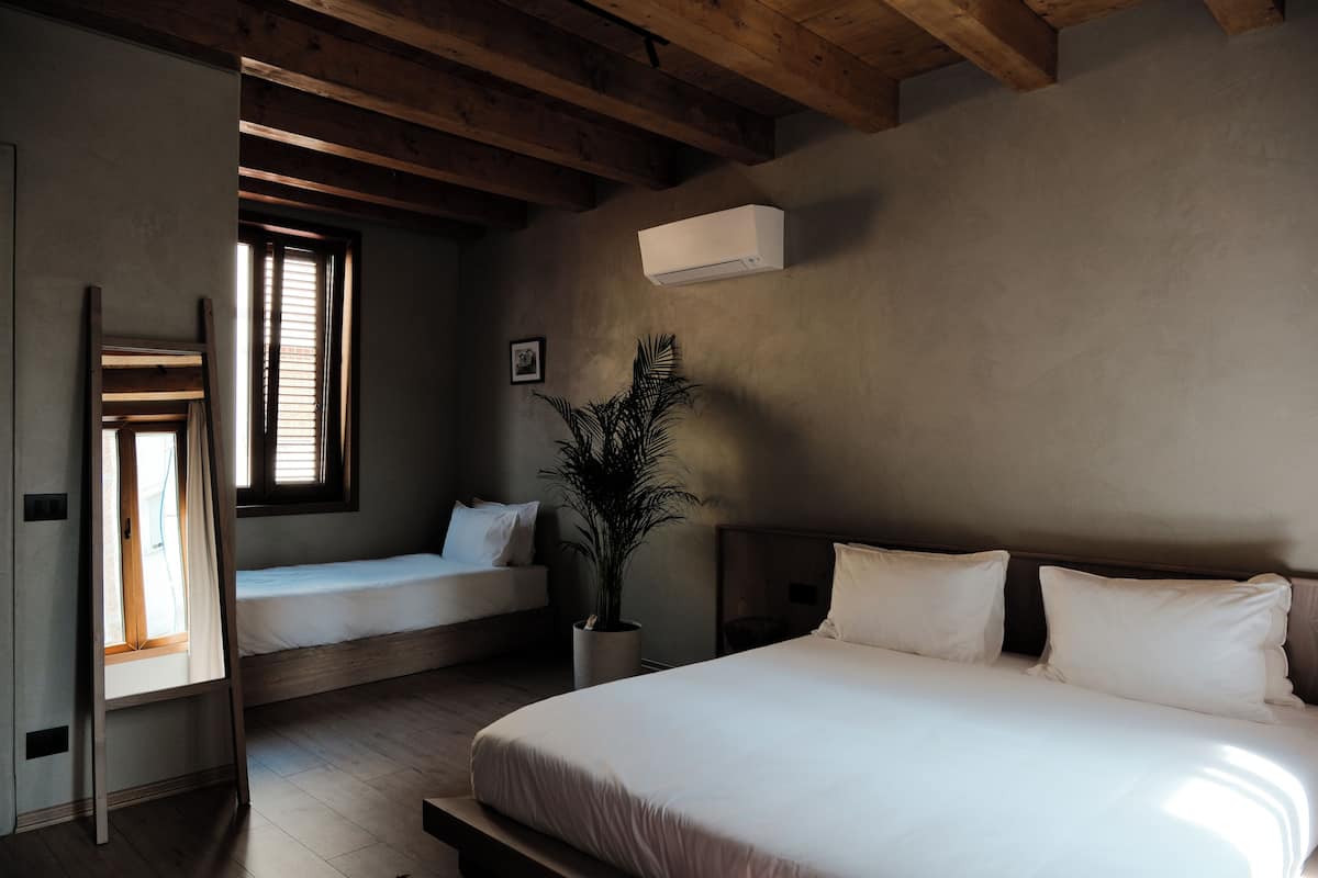 Une chambre à coucher avec un grand lit, un plus petit lit, une plante en pot et une fenêtre avec des stores.
