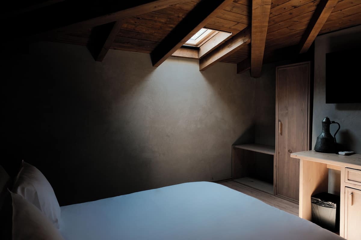 Une chambre avec une lucarne, des poutres en bois, un lit, une table de chevet et une armoire.