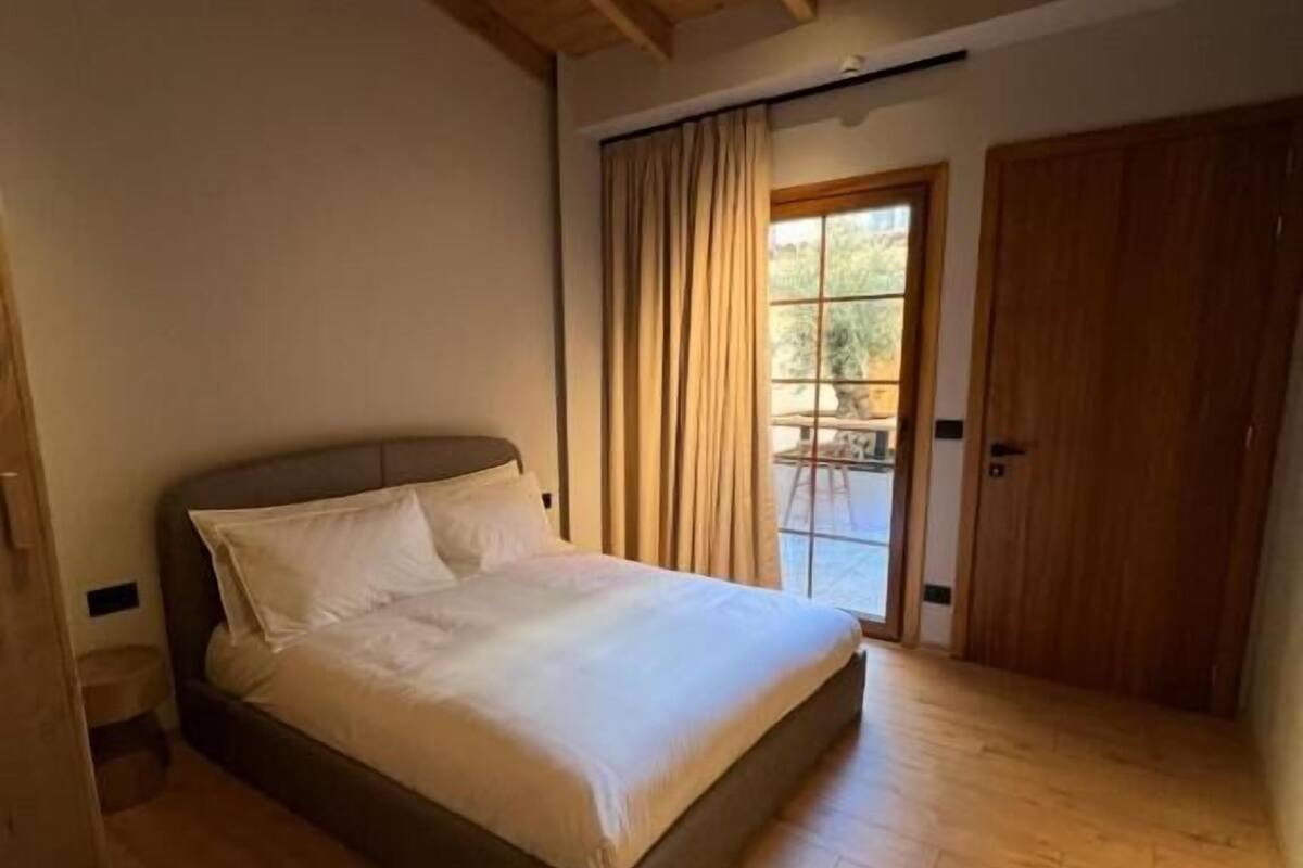 Une chambre avec un plancher en bois, un lit recouvert de draps blancs, un plafond en bois et une porte donnant sur l’extérieur.